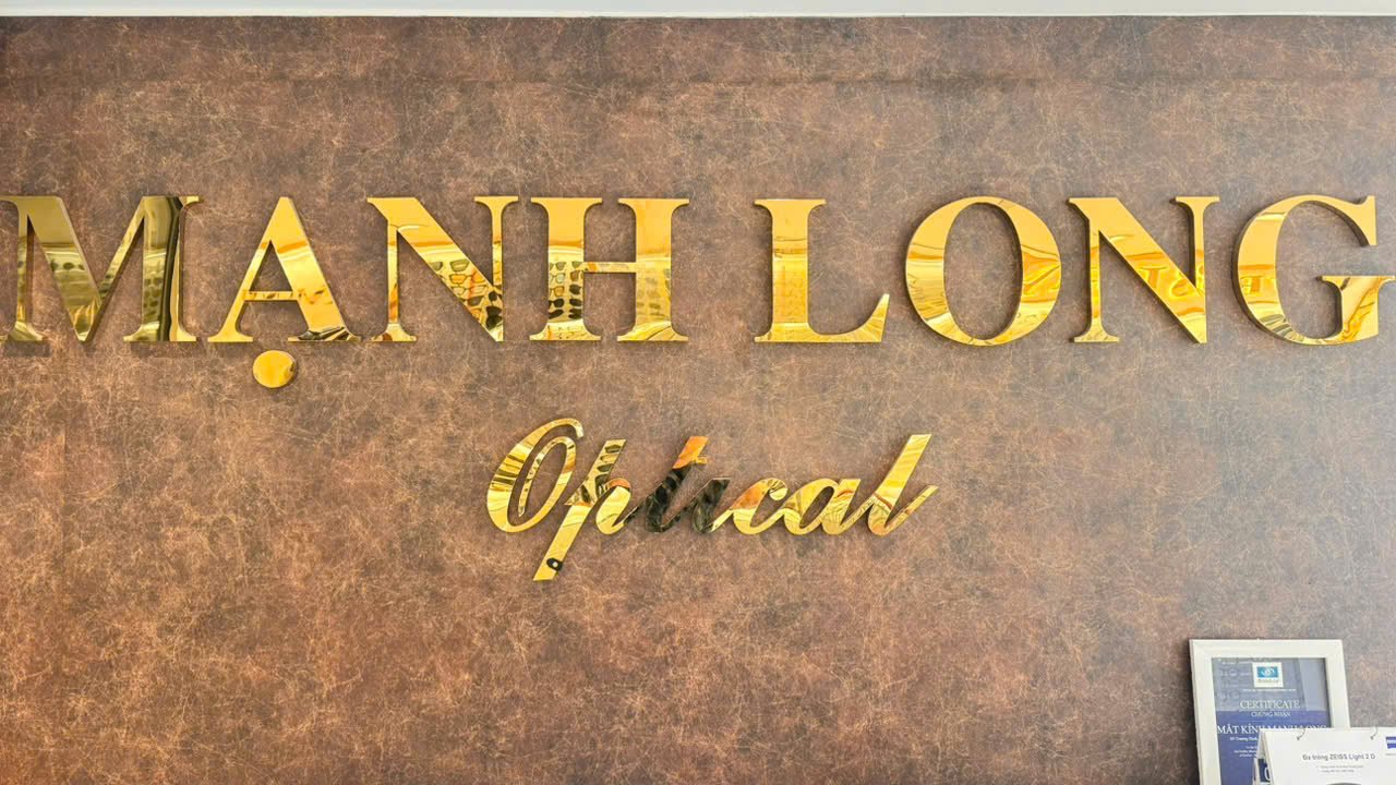 Biển chữ Mạnh Long Optical mạ vàng trên tường giả da nâu tại cửa hàng 147A Lê Duẩn, góc dưới có khung giấy chứng nhận