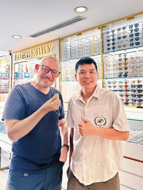 A visitor standing with a Manh Long Optical staff member in front of the frame cabinets at 147A Le Duan, Cua Nam, Hanoi