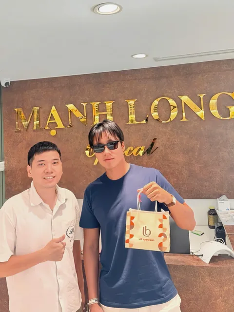 A customer wearing sunglasses, holding a paper bag, standing with a staff member under the Manh Long Optical sign at 147A Le Duan, Cua Nam, Hanoi